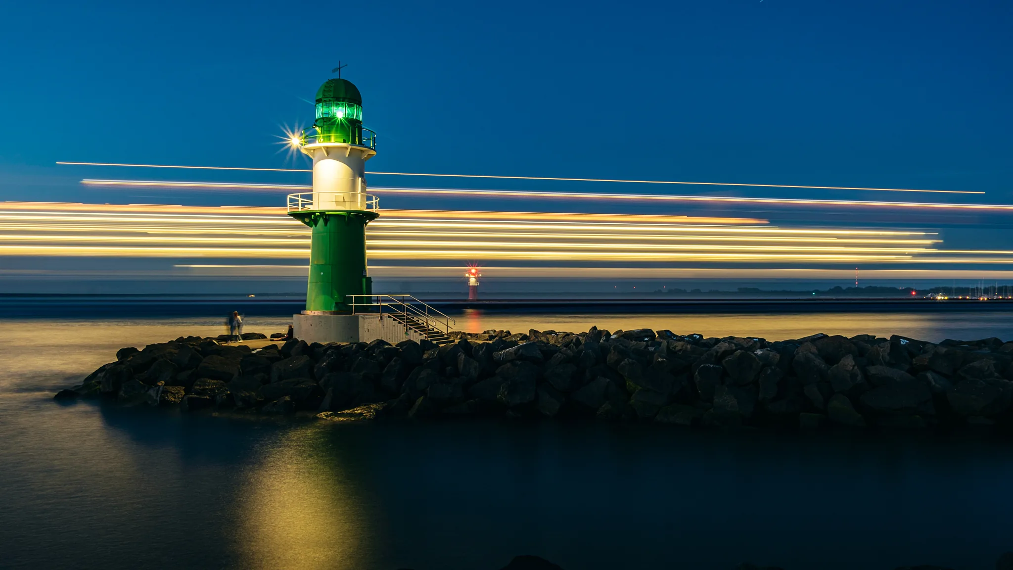 Das grüne Molenfeuer der Westmole in Warnemünde zur blauen Stunde, davor ziehen die Lichtspuren eines auslaufenden Schiffes waagerecht über das Wasser.