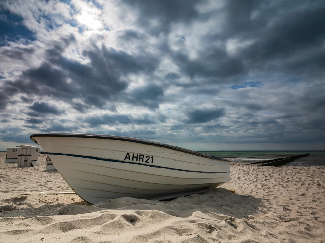 Fischerboot AHR21 am Ostseestrand unter aufziehender Bewölkung, dahinter Strandkörbe und Buhnen.